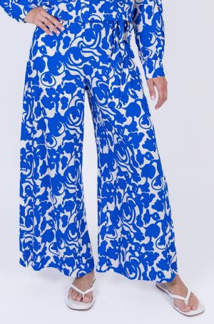 Pantalón Chiringuito de Lylu Wear en blanco con estampado floral azul klein, corte fluido con perneras anchas, cintura elástica y lazada delantera.
